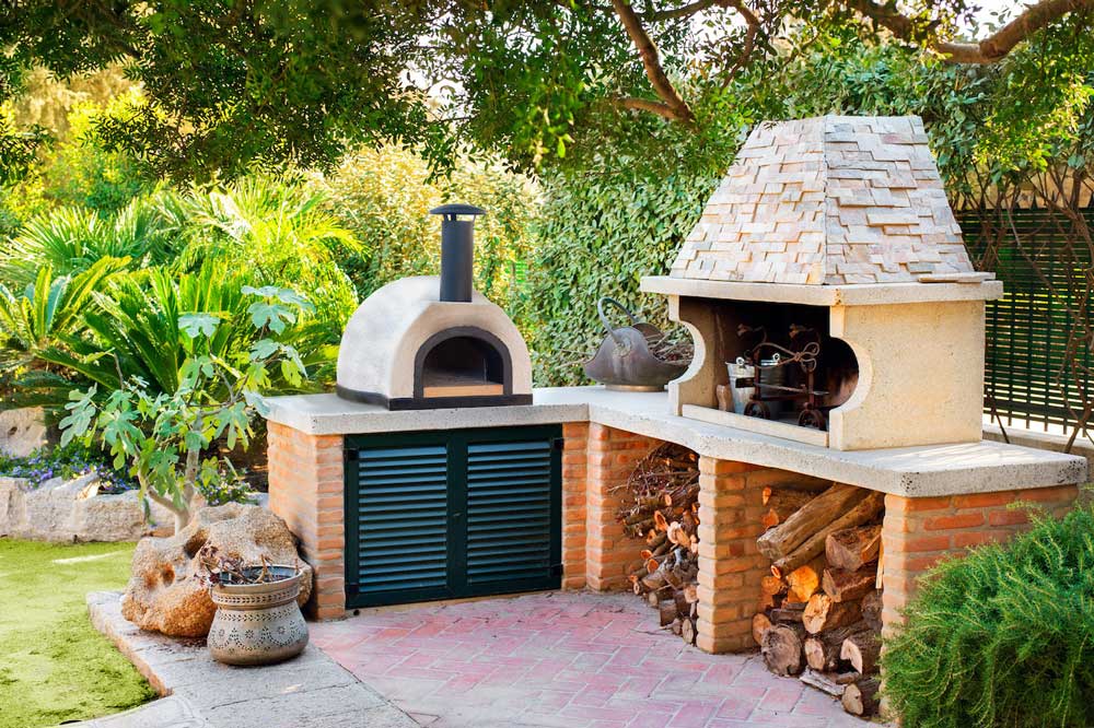 Forno Piombi Santino pizza oven on countertop of an outdoor kitchen. Patio with firewood and landscaping